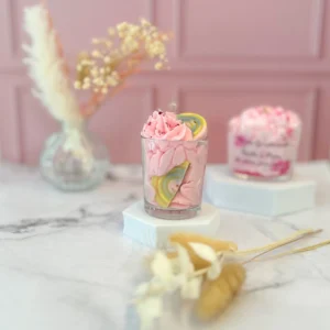 Bougie gourmande artisanale petit modèle dans un verre transparent, avec cire rose fouettée façon chantilly, décorée d’un bonbon arc-en-ciel et de paillettes, posée sur socle blanc dans un décor rose poudré.