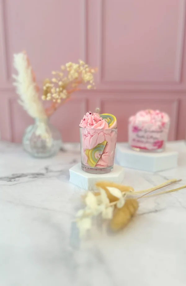 Bougie gourmande artisanale petit modèle dans un verre transparent, avec cire rose fouettée façon chantilly, décorée d’un bonbon arc-en-ciel et de paillettes, posée sur socle blanc dans un décor rose poudré.