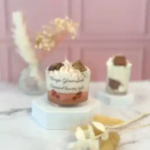 Bougie gourmande au caramel beurre salé dans un grand pot en verre, décorée de crème fouettée en cire et de morceaux façon chocolat, posée sur un socle blanc devant un fond rose.