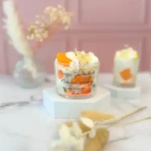 Bougie gourmande dans un grand pot en verre, décorée de crème fouettée blanche et de tranches de clémentine en cire, posée sur un socle blanc avec un fond rose.