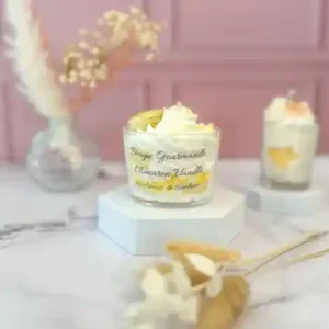Bougie gourmande blanche décorée de cire fouettée et d’un macaron jaune en cire, dans un grand pot en verre, posée sur un socle blanc avec un fond rose.