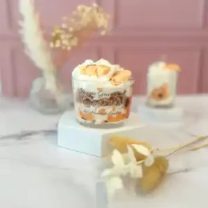 Bougie gourmande dans un grand pot en verre, décorée de cire blanche fouettée et de morceaux façon cookie, posée sur un socle blanc avec un fond rose.