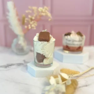 Bougie gourmande à la cire blanche et marron, décorée d’un carré façon caramel en cire, posée sur un socle blanc avec un fond rose.