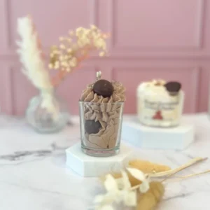 Bougie gourmande à la cire marron, décorée d’une crème fouettée et d’une boule façon truffe en cire, posée sur un socle blanc avec un fond rose.
