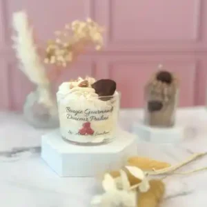 Bougie gourmande blanche dans un grand pot en verre, décorée de crème fouettée en cire et de chocolats en cire, posée sur un socle blanc avec un fond rose.