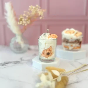 Bougie gourmande avec de la cire blanche fouettée et un biscuit façon cookie, posée sur un socle blanc avec un fond rose.
