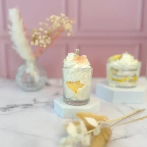 Bougie gourmande décorée de cire blanche fouettée et d’un macaron jaune, posée sur un support blanc avec un fond rose.