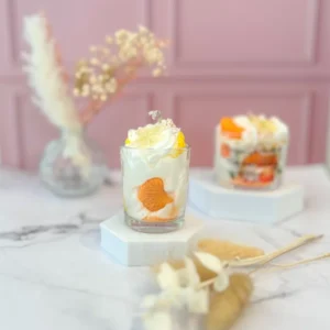 Bougie gourmande à la cire blanche, décorée d’une tranche de clémentine en cire, posée sur un socle blanc avec un fond rose.