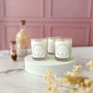 Trois bougies artisanales blanches en verre transparent, alignées sur un socle blanc, avec étiquette Lez'arômes de Sandrine, sur fond rose poudré et éléments déco floraux naturels.
