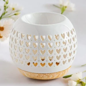 Brûle-parfum Coeur blanc en céramique ajourée de petits cœurs, avec base en bois clair, entouré de fleurs blanches.