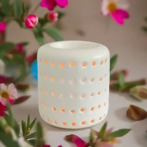 Brûle-parfum Coliseo cylindrique blanc ajouré de petits trous ronds, allumé, entouré de fleurs roses.