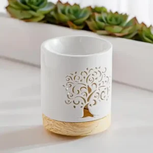 Brûle-parfum en céramique blanche avec un motif ajouré en forme d'arbre de vie, sur une base en bois clair, posé devant des plantes grasses.