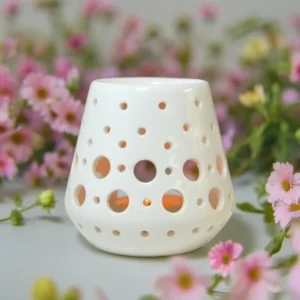 Brûle-parfum Loob blanc en céramique ajourée avec motifs circulaires, posé devant un fond de fleurs roses.