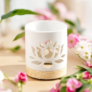 Brûle-parfum blanc en céramique avec motif nénuphar découpé, posé sur une base en bois clair, entouré de fleurs roses et blanches.
