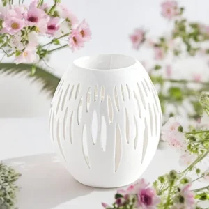 Brûle-parfum blanc en forme d’ovale, ajouré de motifs organiques, entouré de fleurs roses sur fond blanc.