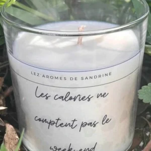 Bougie blanche en pot transparent avec l’inscription "Les calories ne comptent pas le week-end", posée dans l’herbe.