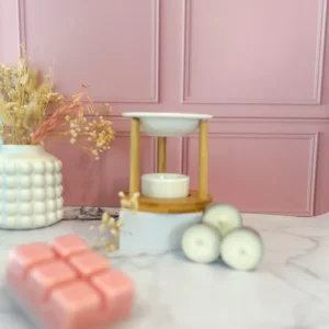 Brûleur de fondants en céramique blanche et bois, au design épuré et moderne, accompagné de bougies chauffe-plat et de fondants parfumés roses, sur fond rose pastel et marbre clair.