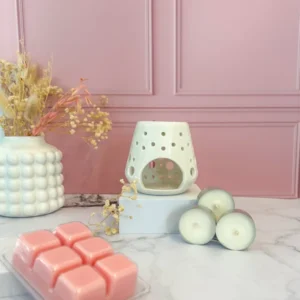 Brûleur de fondants en céramique blanche perforée, accompagné de bougies chauffe-plat et d’un pack de fondants parfumés roses, sur fond rose élégant et marbre clair.
