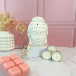 Brûleur de fondants en forme de tête de Bouddha blanc, accompagné de bougies chauffe-plat et d’un pack de fondants parfumés roses, dans une ambiance zen sur fond rose poudré.