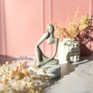 Sculpture artisanale en Jesmonite grise représentant une silhouette féminine assise en position méditative, posée sur un socle blanc, avec fleurs séchées et mur rose poudré en arrière-plan.
