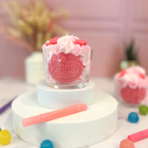 Bougie artisanale rose et blanche “Merci Maîtresse” en cire végétale, posée sur un socle rond blanc, avec un décor floral et un fond rose doux.