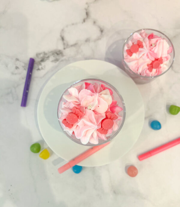 Vue du dessus de deux bougies gourmandes “Merci Maîtresse” en cire végétale rose et blanche, entourées de stylos et de petits bonbons.